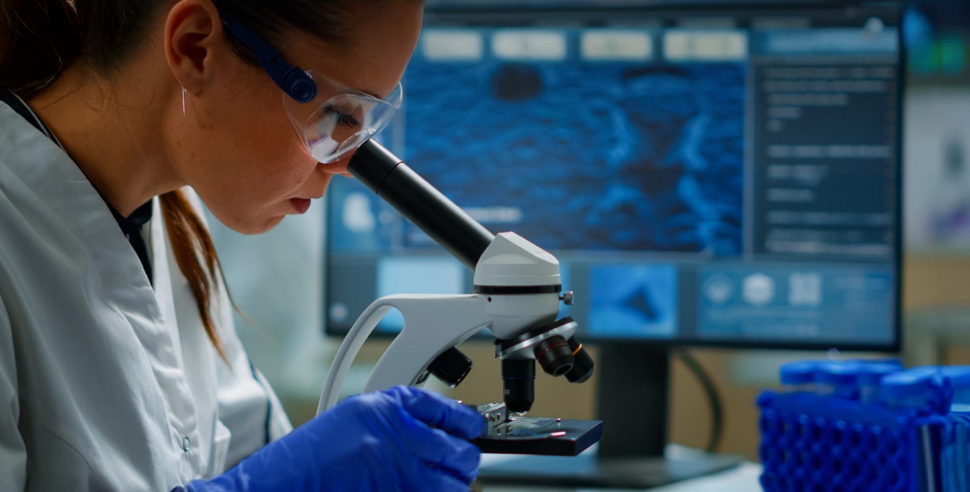 A researcher in a lab coat and safety glasses examining a sample under a microscope